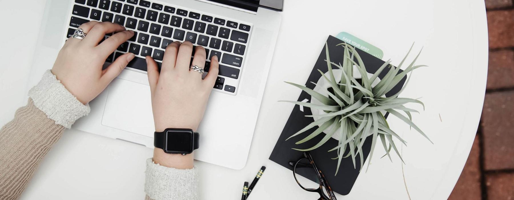 overhead shot of woman working on a laptop at a round table with pencils, her cell phone, glasses, notebook and a potted plant